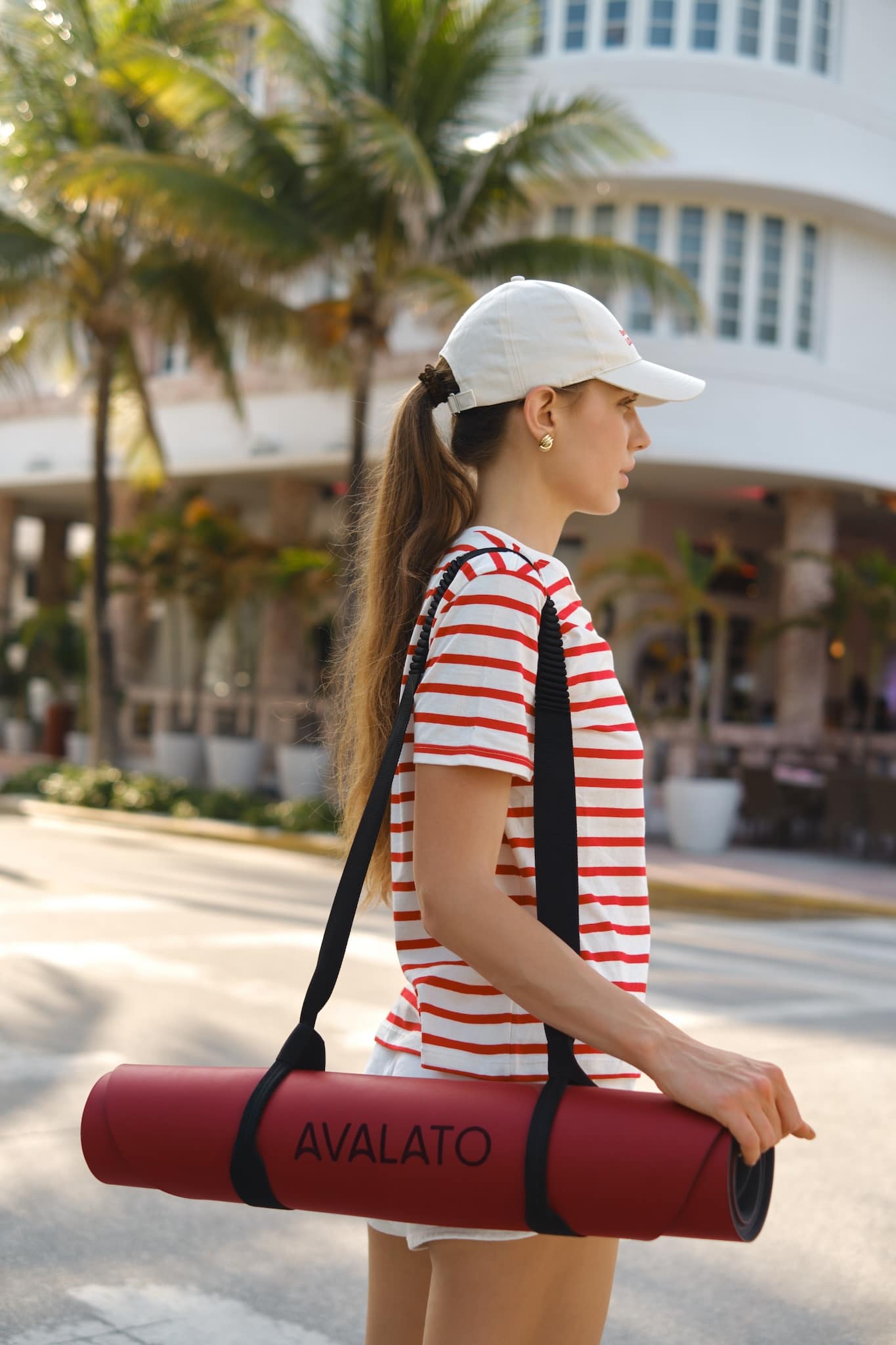 Athletic woman carrying a red AVALATO exercise mat on Miami beach with palm trees in the background