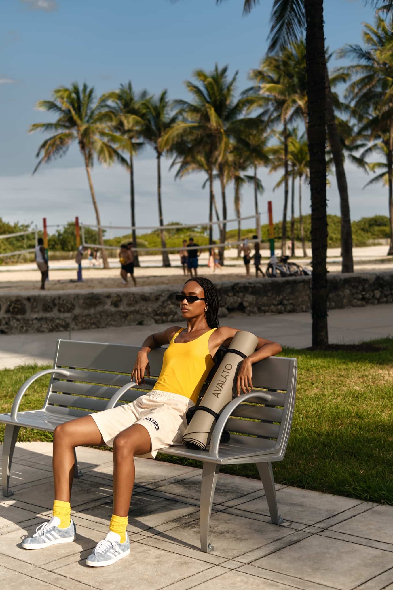 Athletic woman, relaxing on a bench with a beige exercise mat in a miami beach park with palm trees and a beach volleyball court in the background.