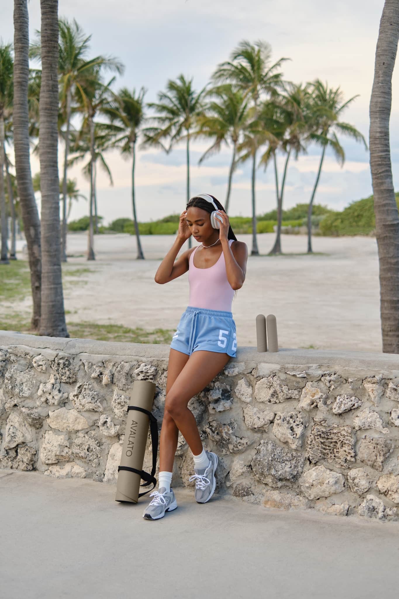 Athletic woman wearing fitness outfit, with AVALATO workout gear including an exercise mat and silicone dumbbells, relaxing by a stone wall near the beach