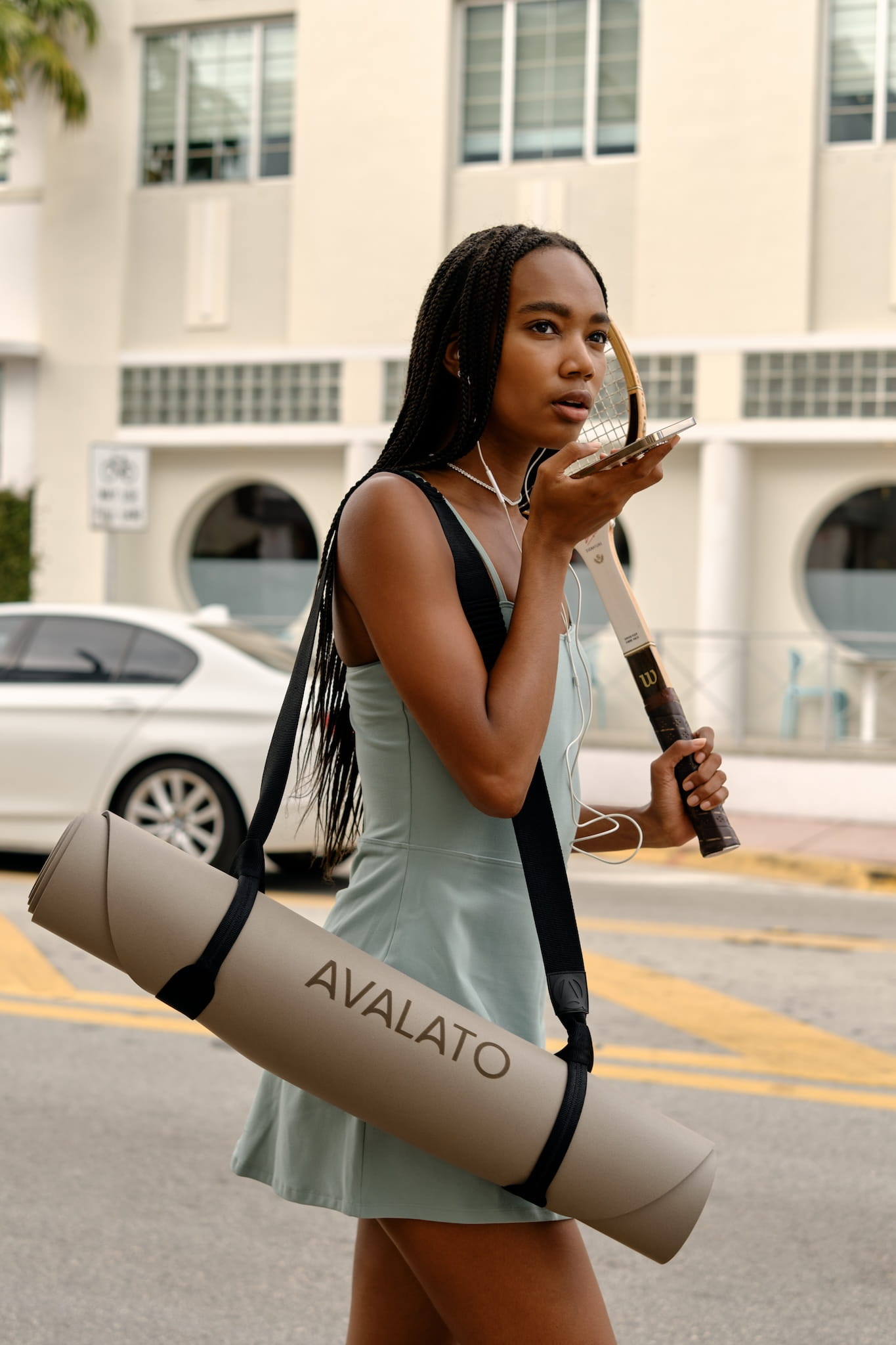 Athletic woman, carrying a beige yoga mat and tennis racket, speaking on a smartphone, walking in an urban area with buildings in the background