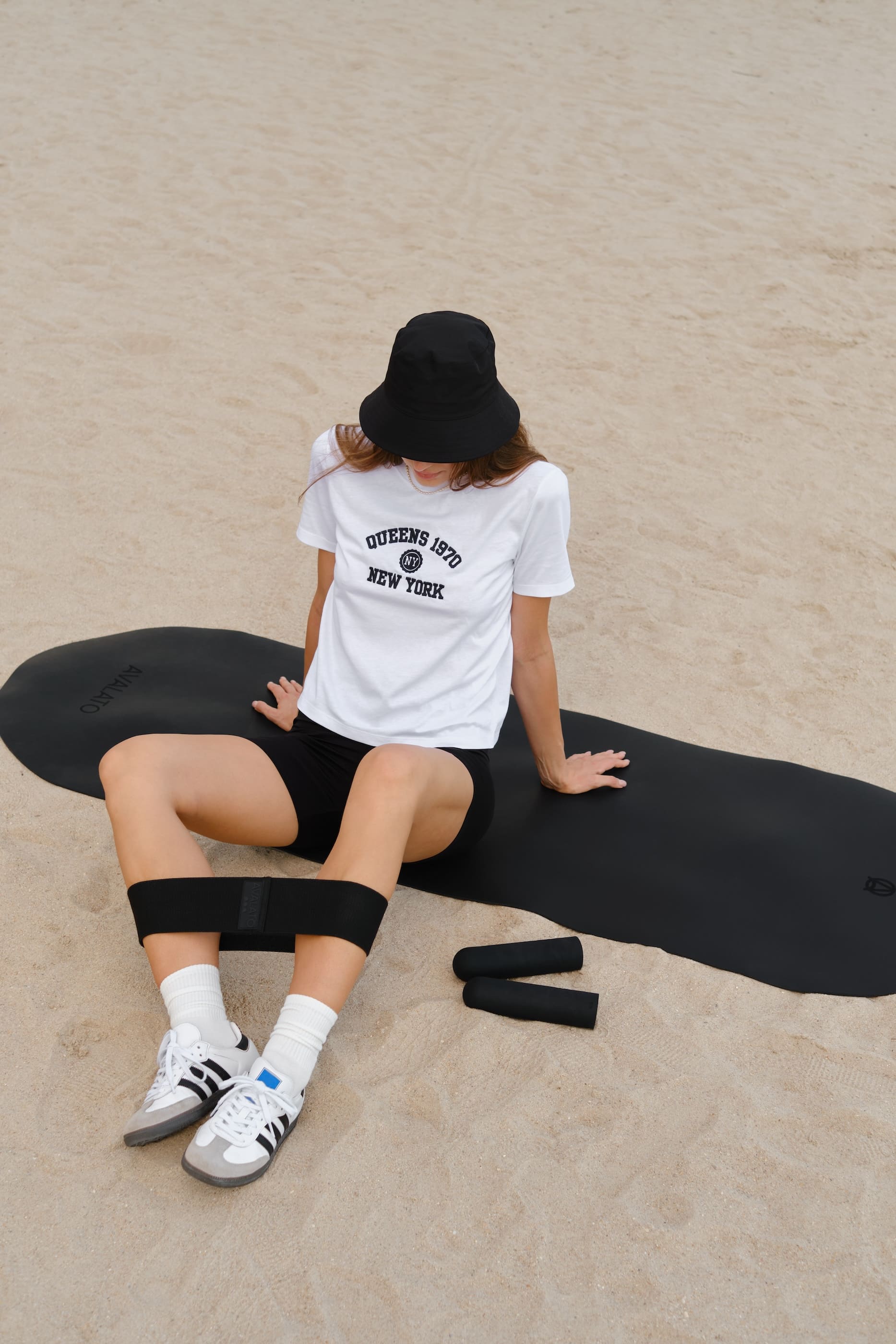 Woman sitting on AVALATO black exercise mat on the beach with resistance bands and silicone dumbbells