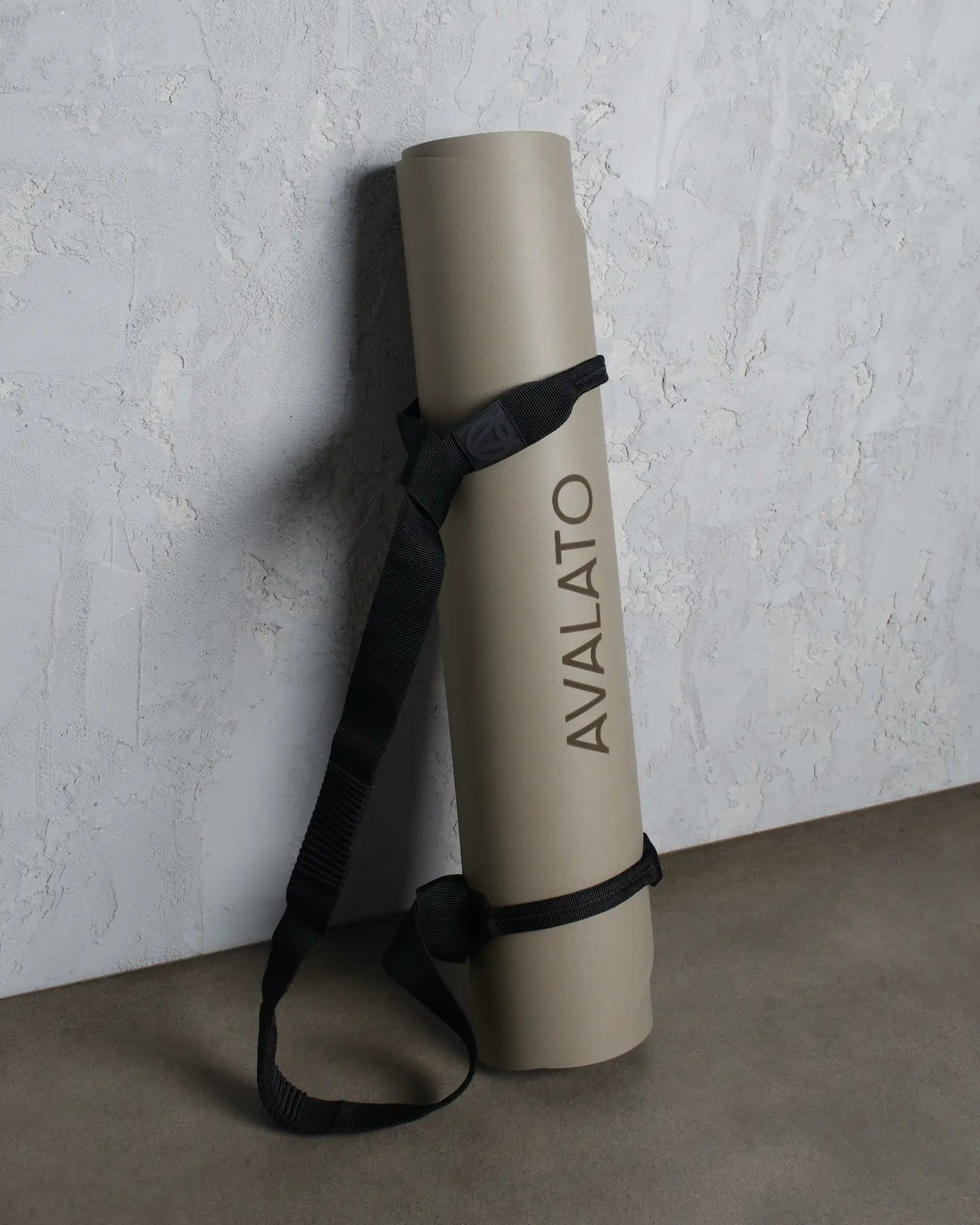 Beige avalato exercise mat with carrying yoga strap, leaning against a textured wall