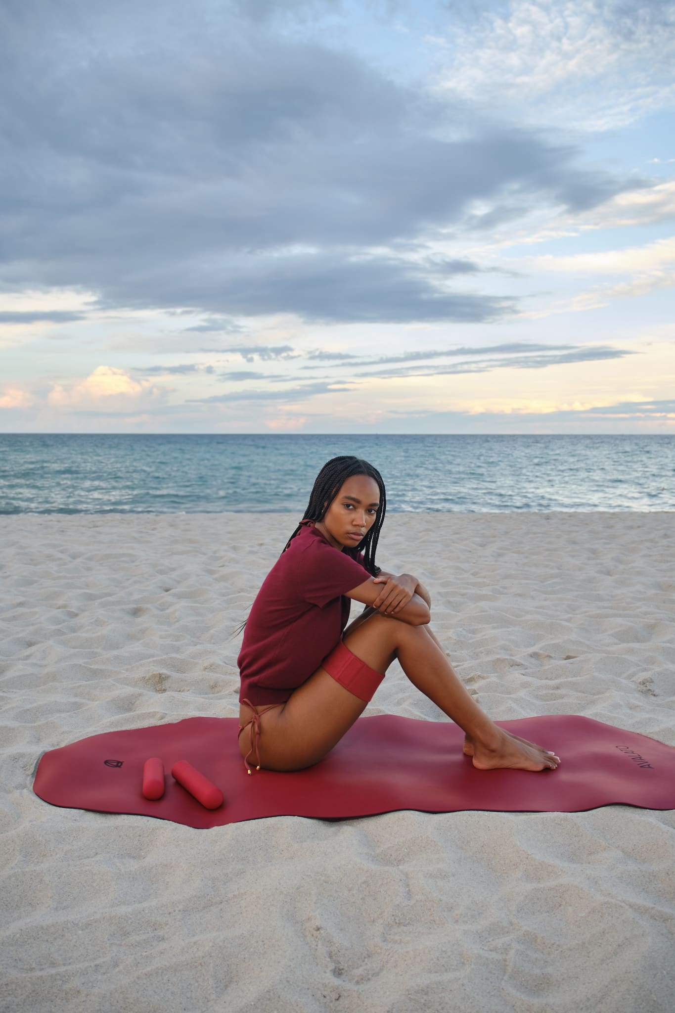 Fit woman wearing a red shirt and bikini swimwear, sitting on a red yoga mat at the beach with silicone dumbbells and resistance band on her legs