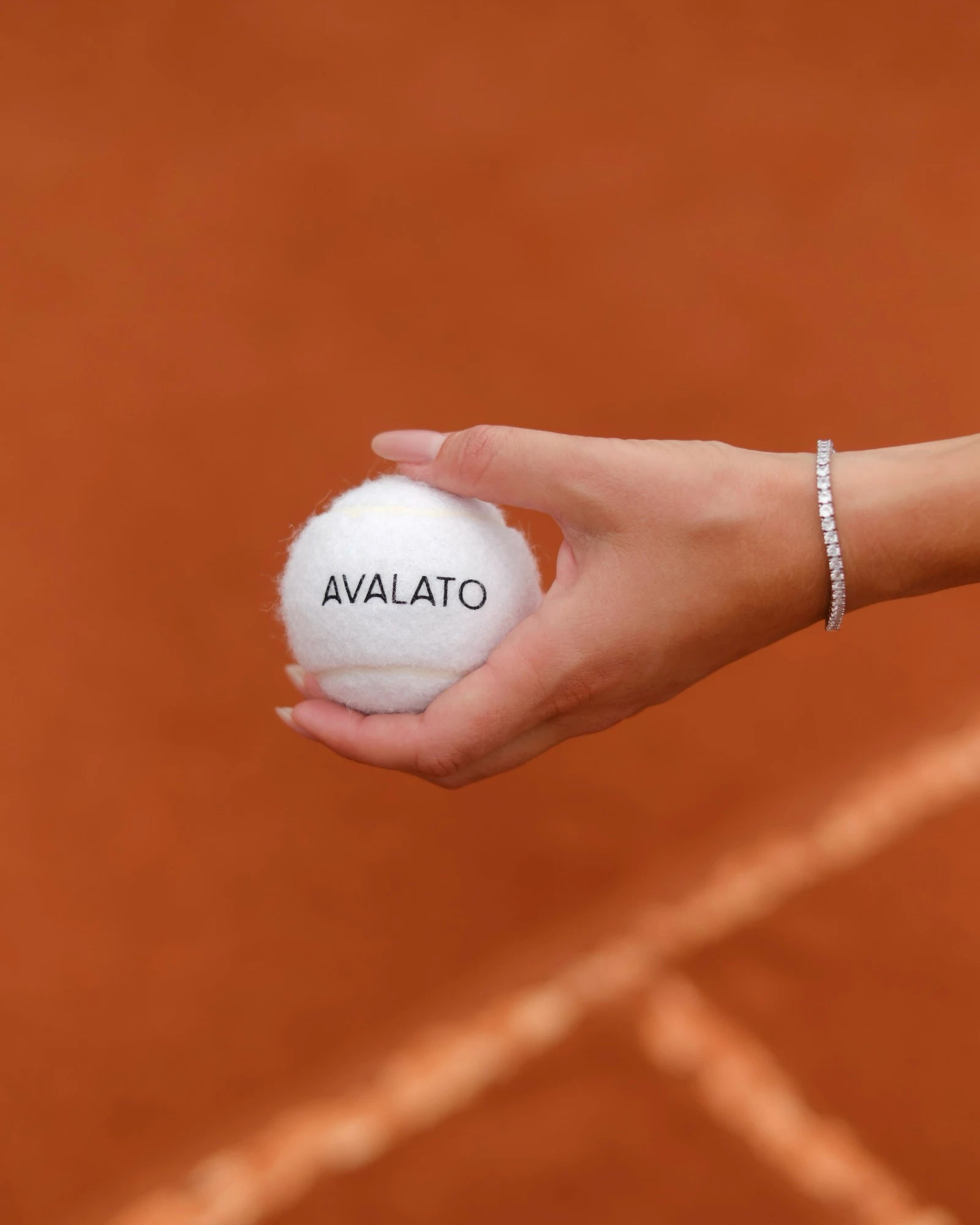 A hand holding an Avalato white tennis ball on a clay court, featuring a tennis aesthetic with vintage style