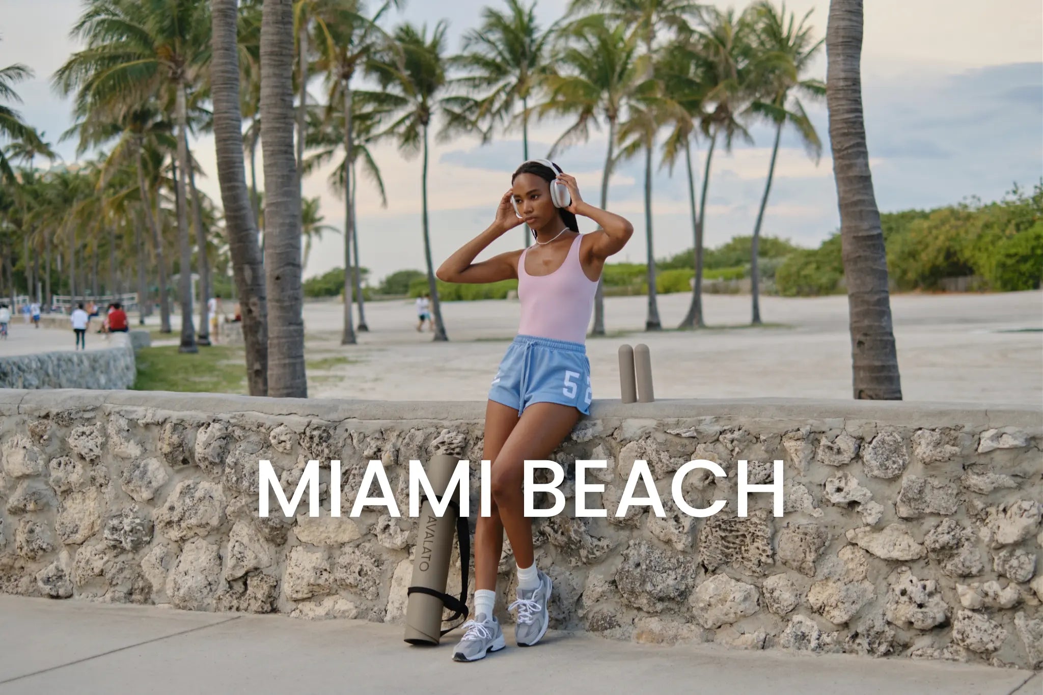 Woman in workout attire standing by a stone wall with headphones, palm trees, and beach in the background, with "Miami Beach" text overlay