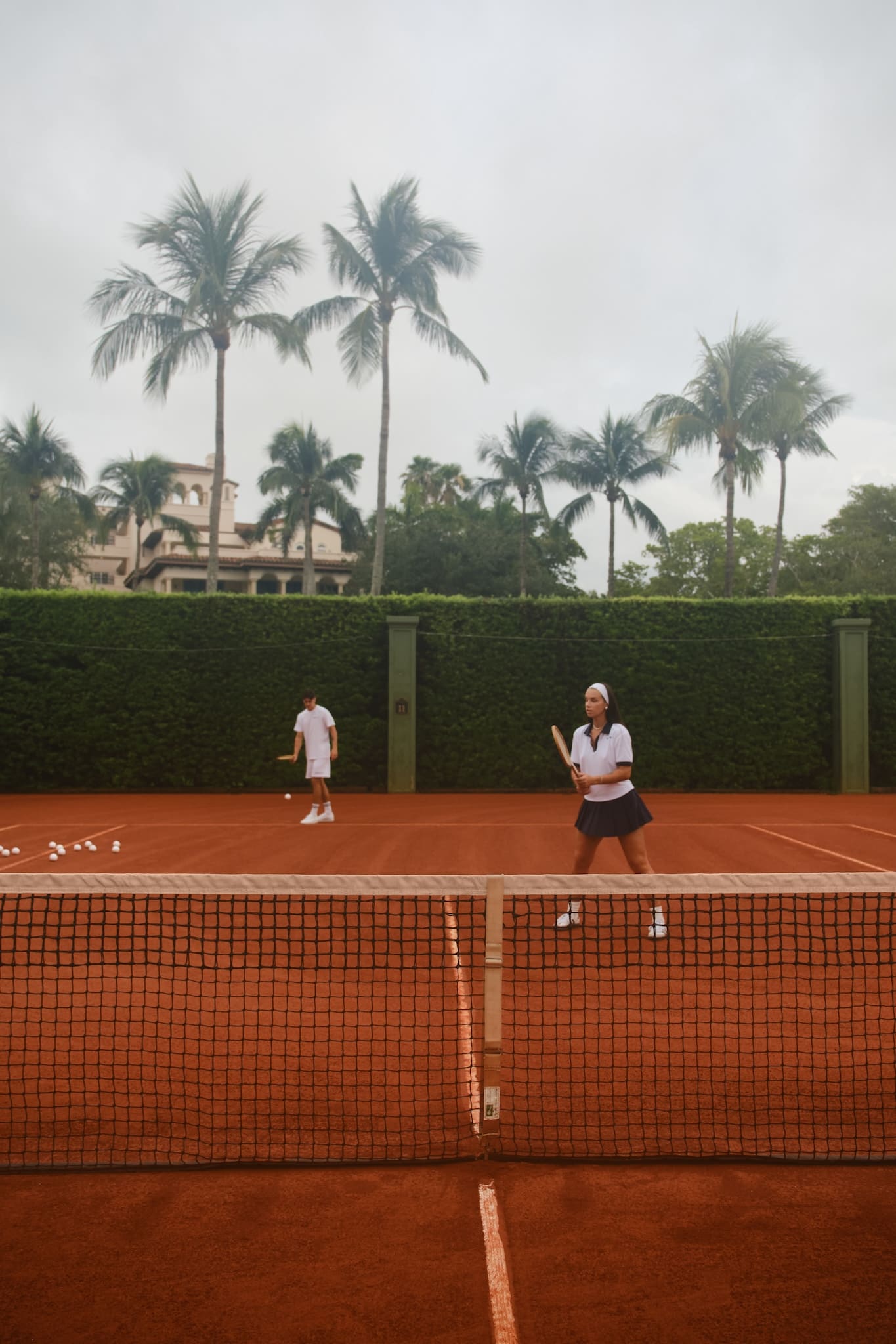 Two tennis players in vintage attire on a clay court, preparing for a match with palm trees and buildings in the background