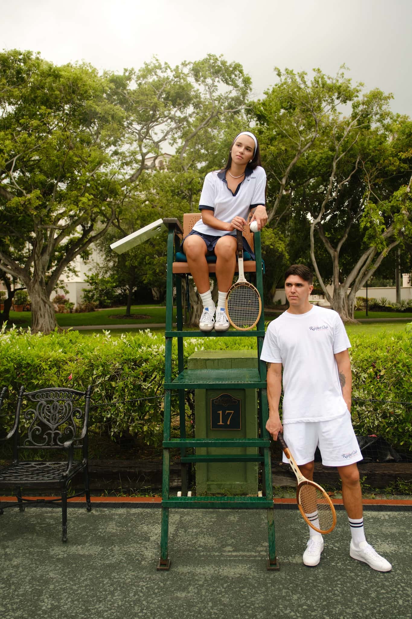 A tennis couple in the racquet club with vintage wooden rackets, one sitting on a chair umpire stand and the other standing beside it on a tennis court