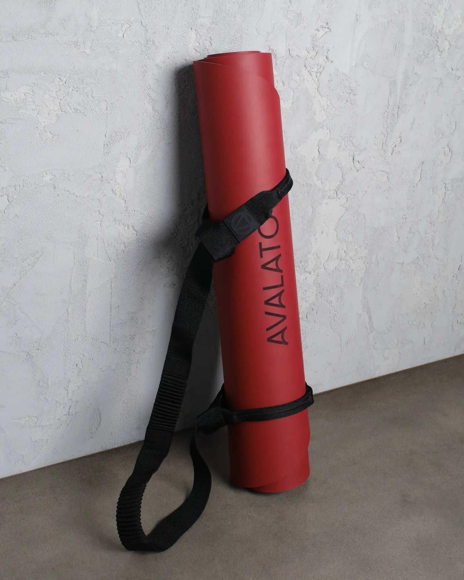 Red avalato exercise mat with carrying yoga strap, leaning against a textured wall