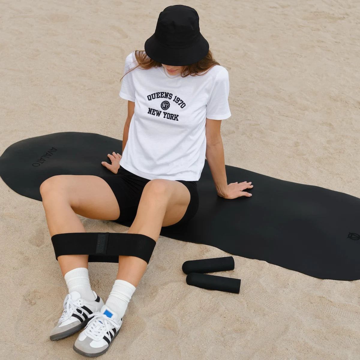 Woman in a white T-shirt and black toga shorts sitting on a black exercise mat at the beach with a black resistance band around her legs and silicone dumbbells on the sand