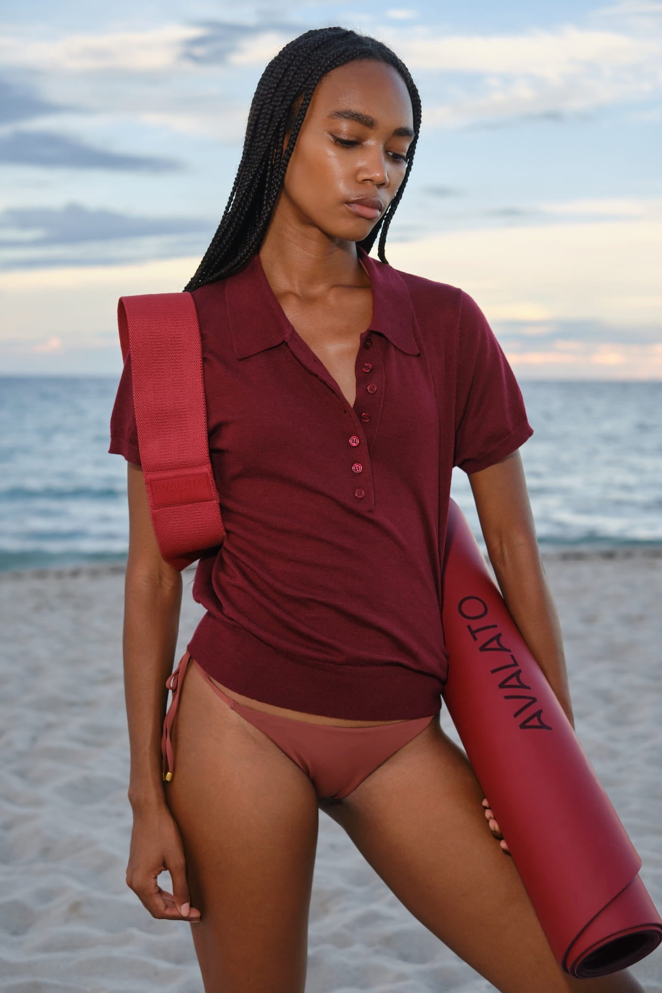 Woman wearing a red shirt and bikini, holding a red AVALATO exercise mat and a red resistance band on the beach at sunset