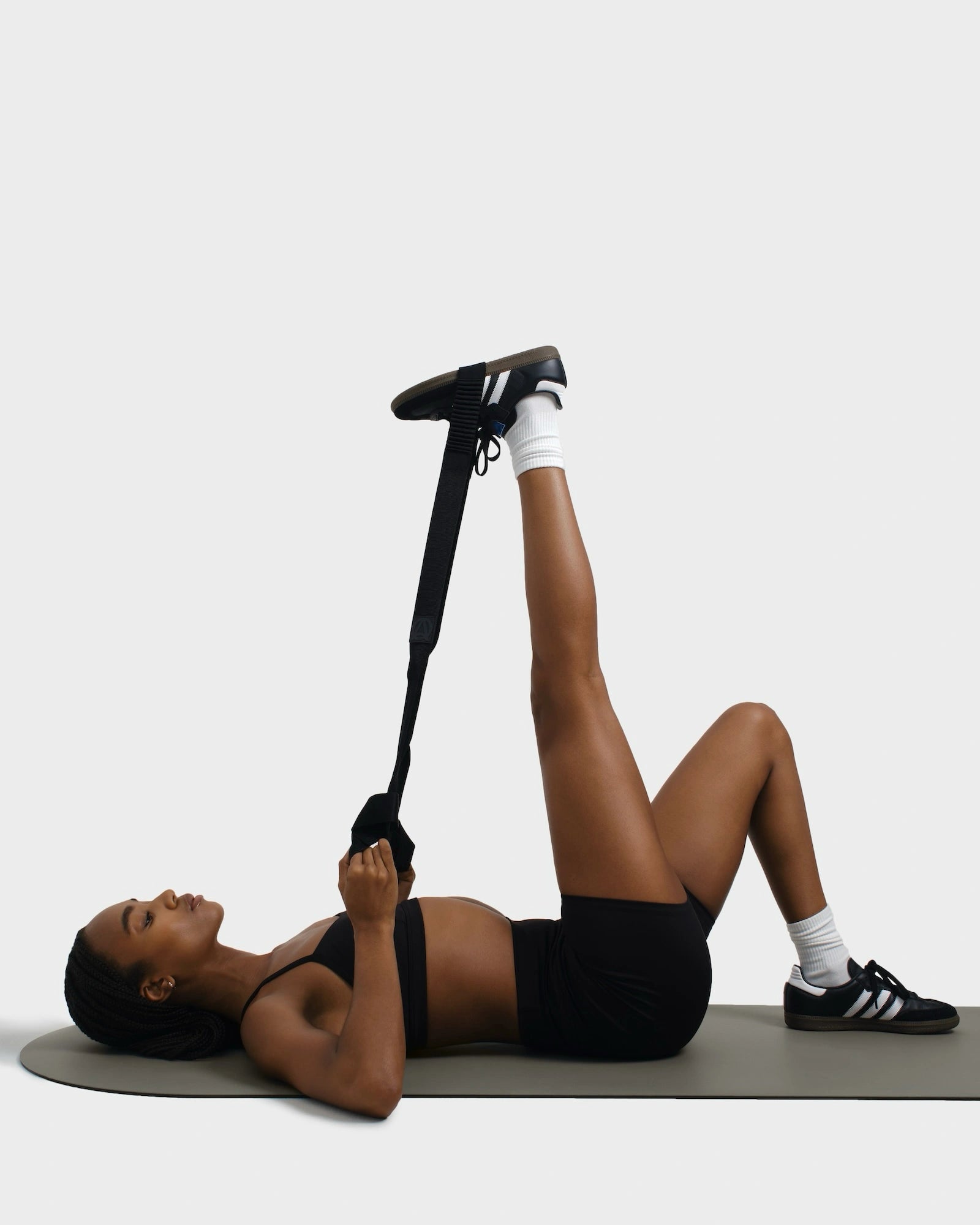 Woman in gym outfit lying on a beige exercise mat, stretching her leg using a black yoga strap