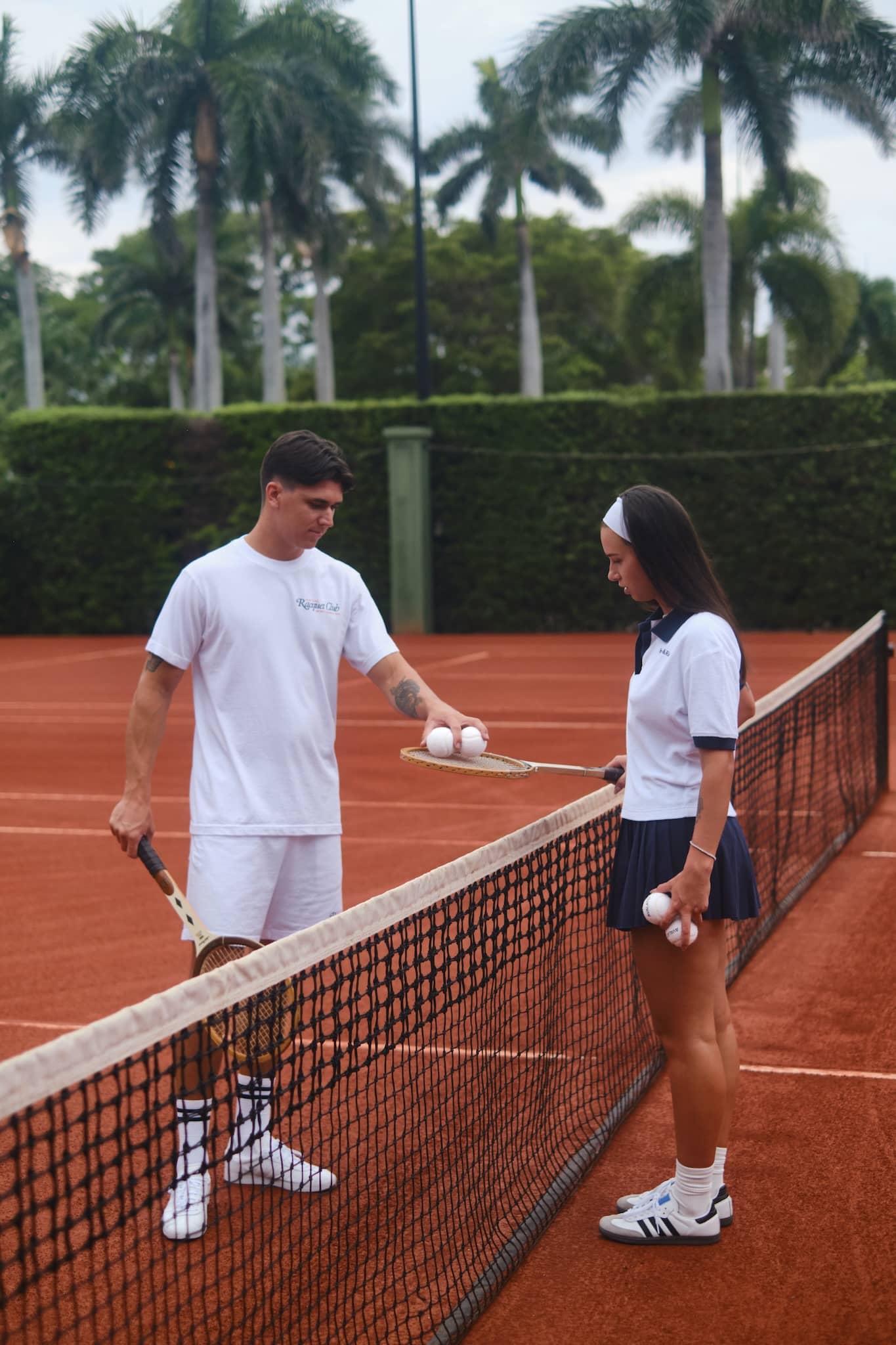 Two tennis players in old money style attire with wooden rackets standing on a clay court