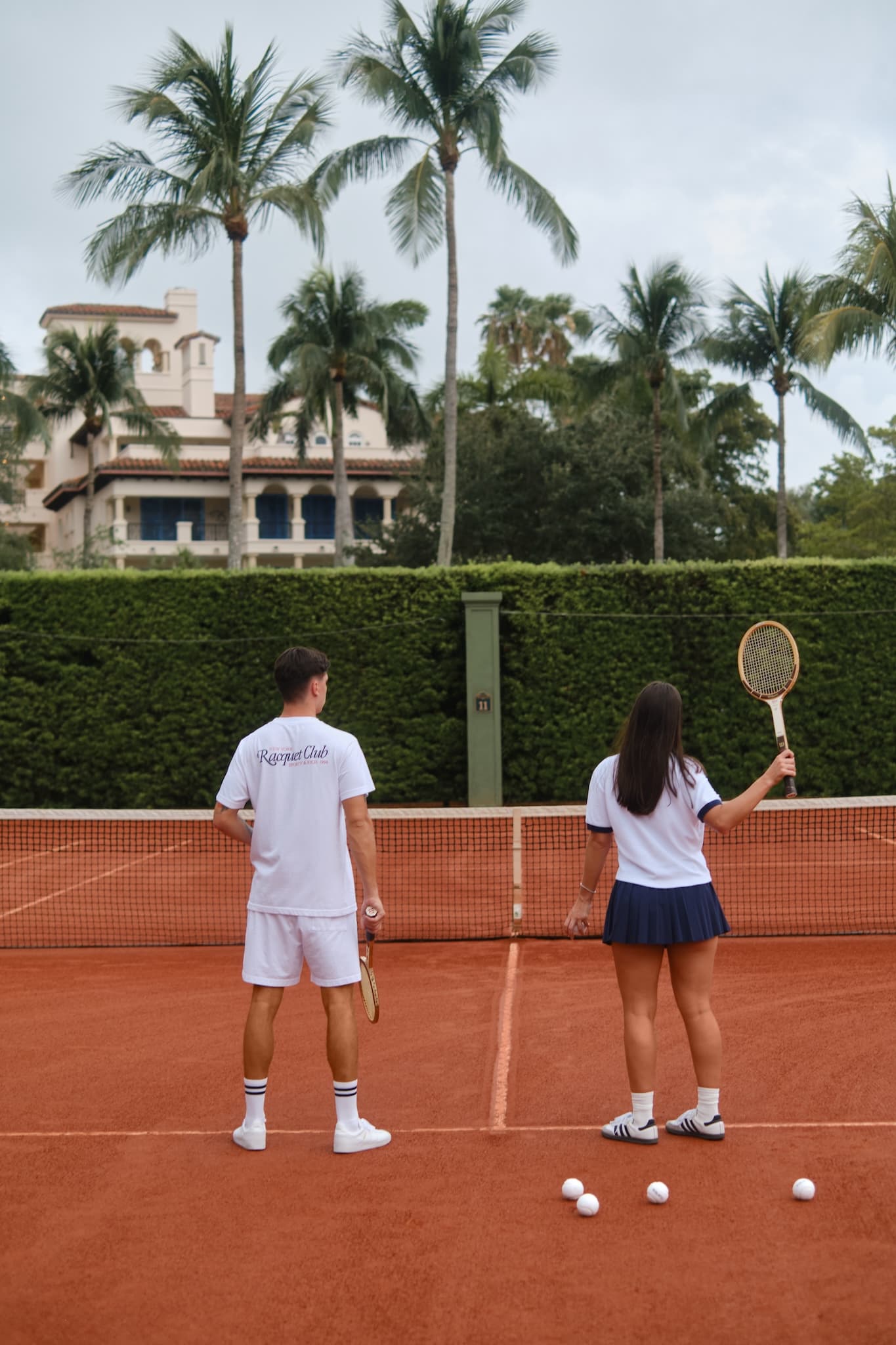 Two tennis players in the racquet club standing on a clay court with their backs to the camera, facing the net