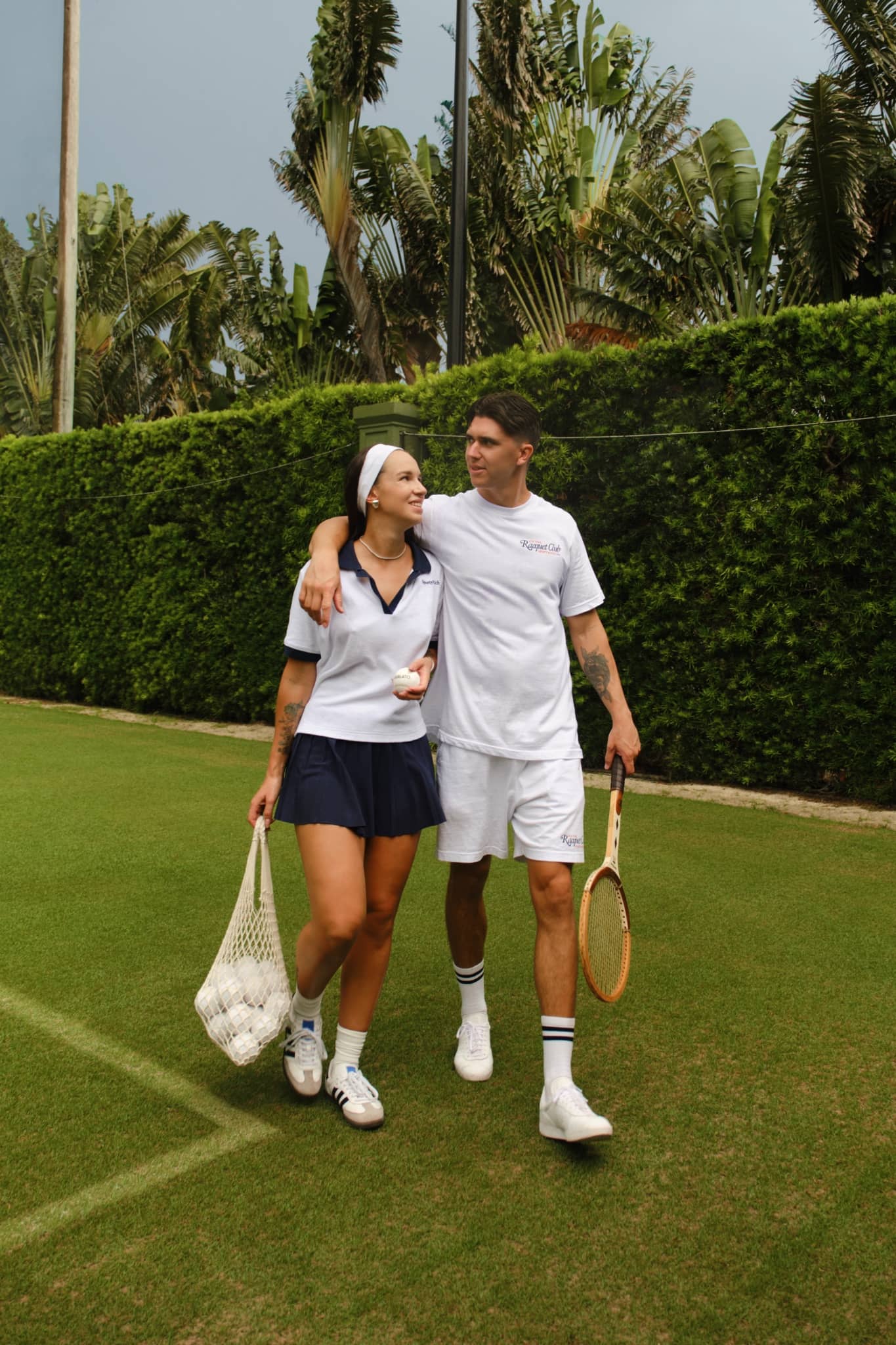 Two tennis players in vintage racquet club walking together on a grass court, carrying a wooden racket and a net bag filled with white tennis balls