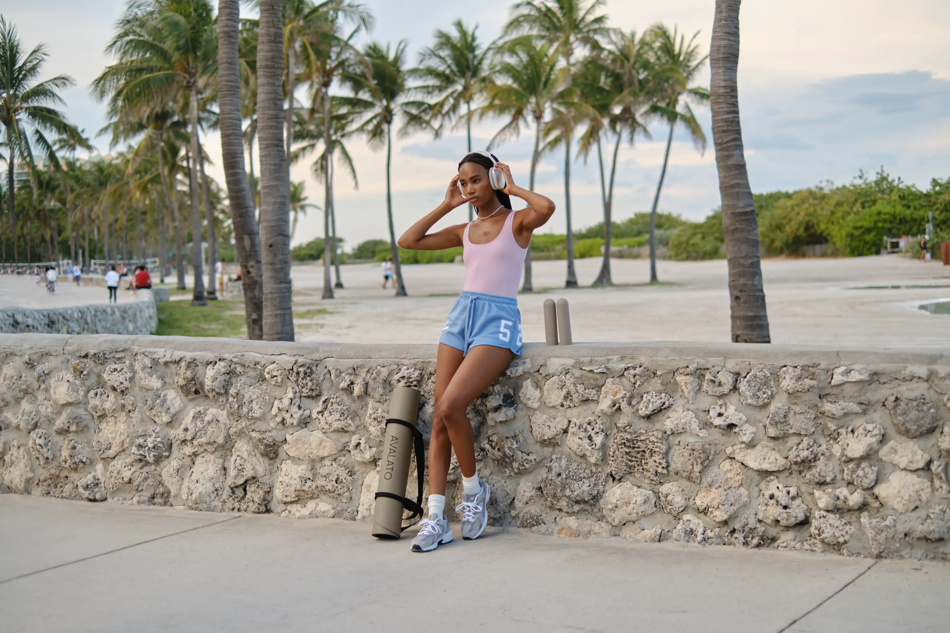 Woman wearing a fitness outfit, relaxing by a stone wall with AVALATO workout gear including an exercise mat and silicone dumbbells at a beach