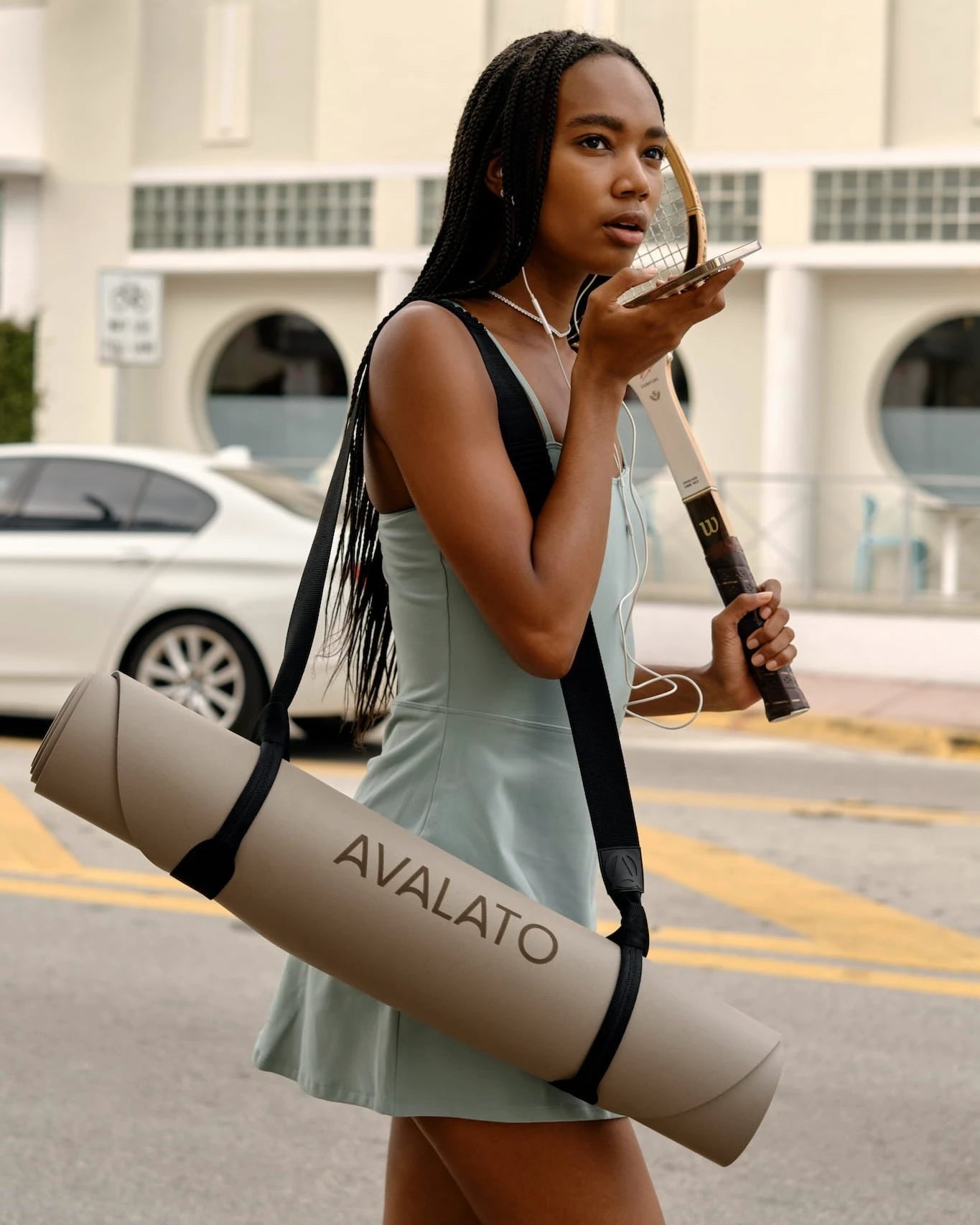 Athletic woman, carrying a beige yoga mat and tennis racket, speaking on a smartphone, walking in an urban area with buildings in the background