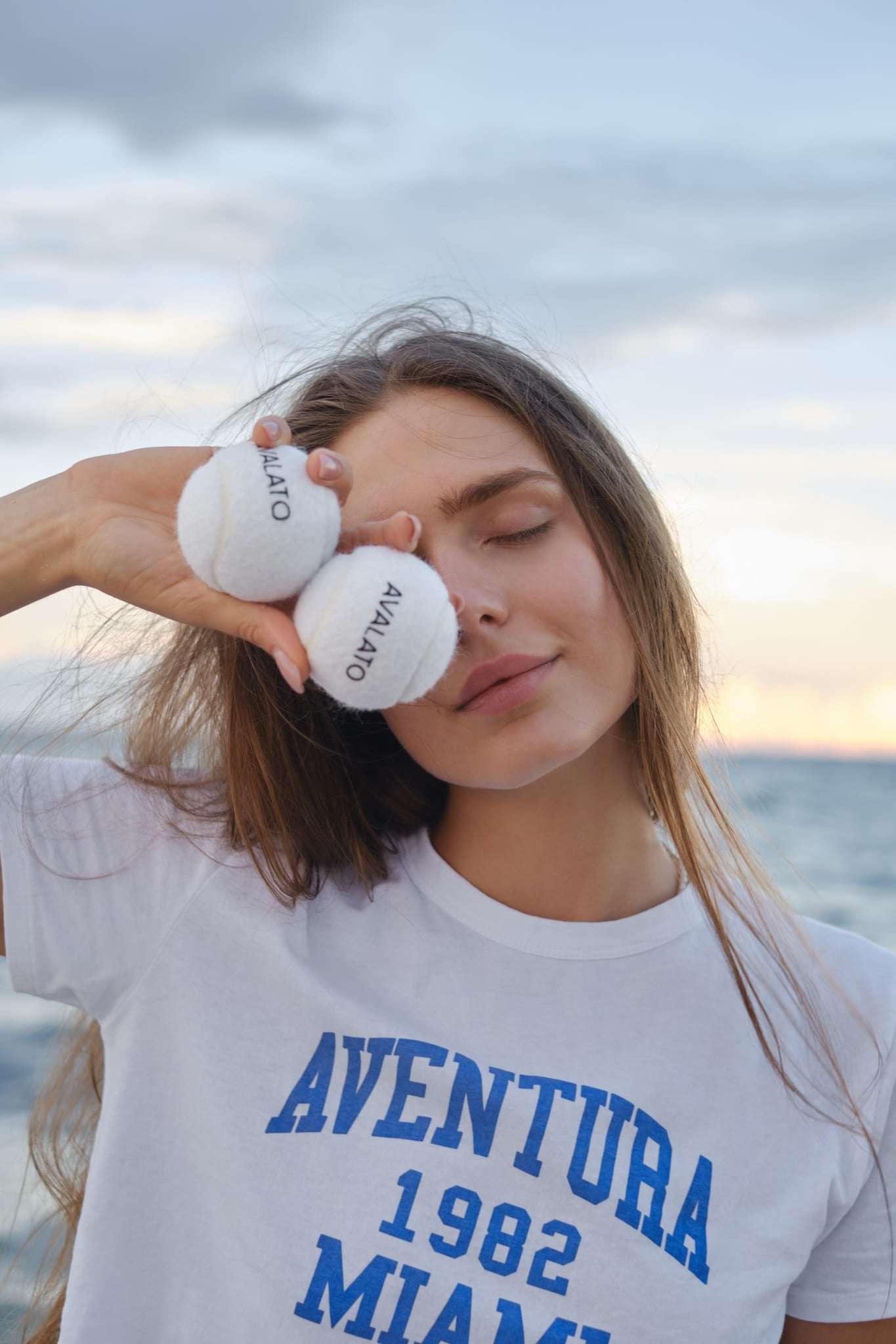 Woman wearing a white activewear, holding two AVALATO tennis balls near her face at the beach with the ocean in the background