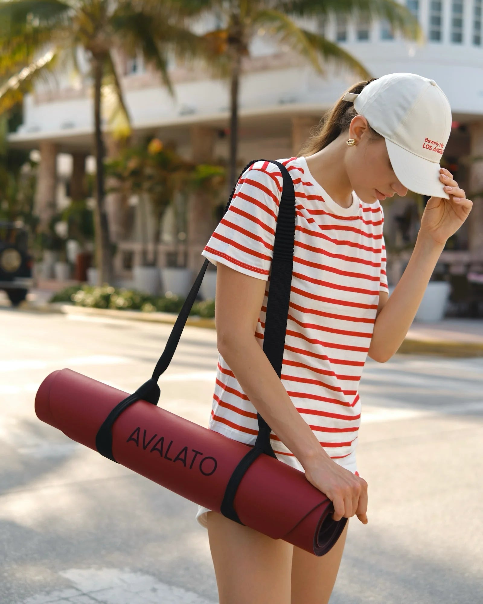 Athletic woman carrying a red AVALATO exercise mat on Miami beach with palm trees in the background